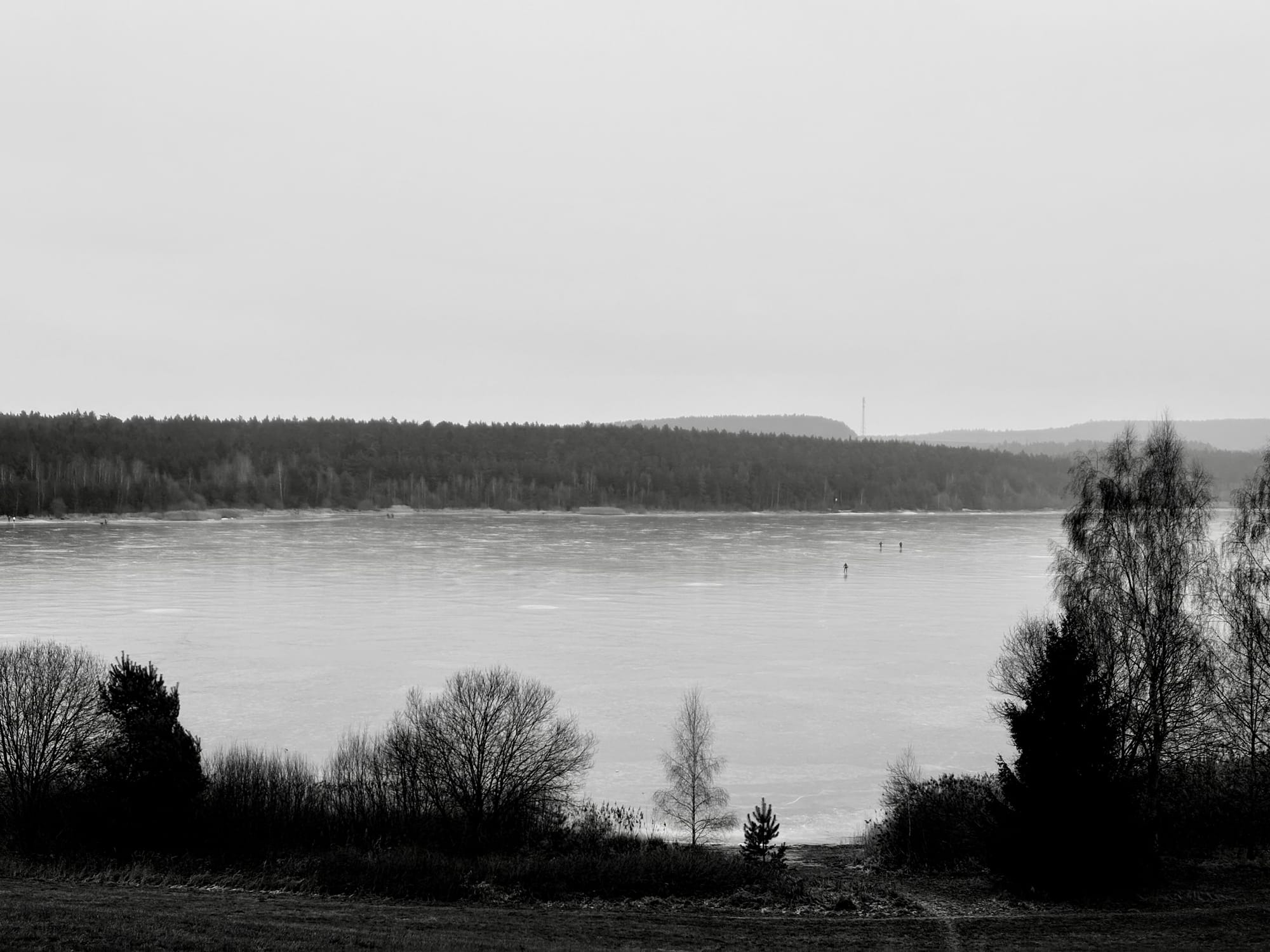 Ein See, zugefroren. Das Foto ist schwarz-weiß. Menschen laufen darauf Schlittschuh.