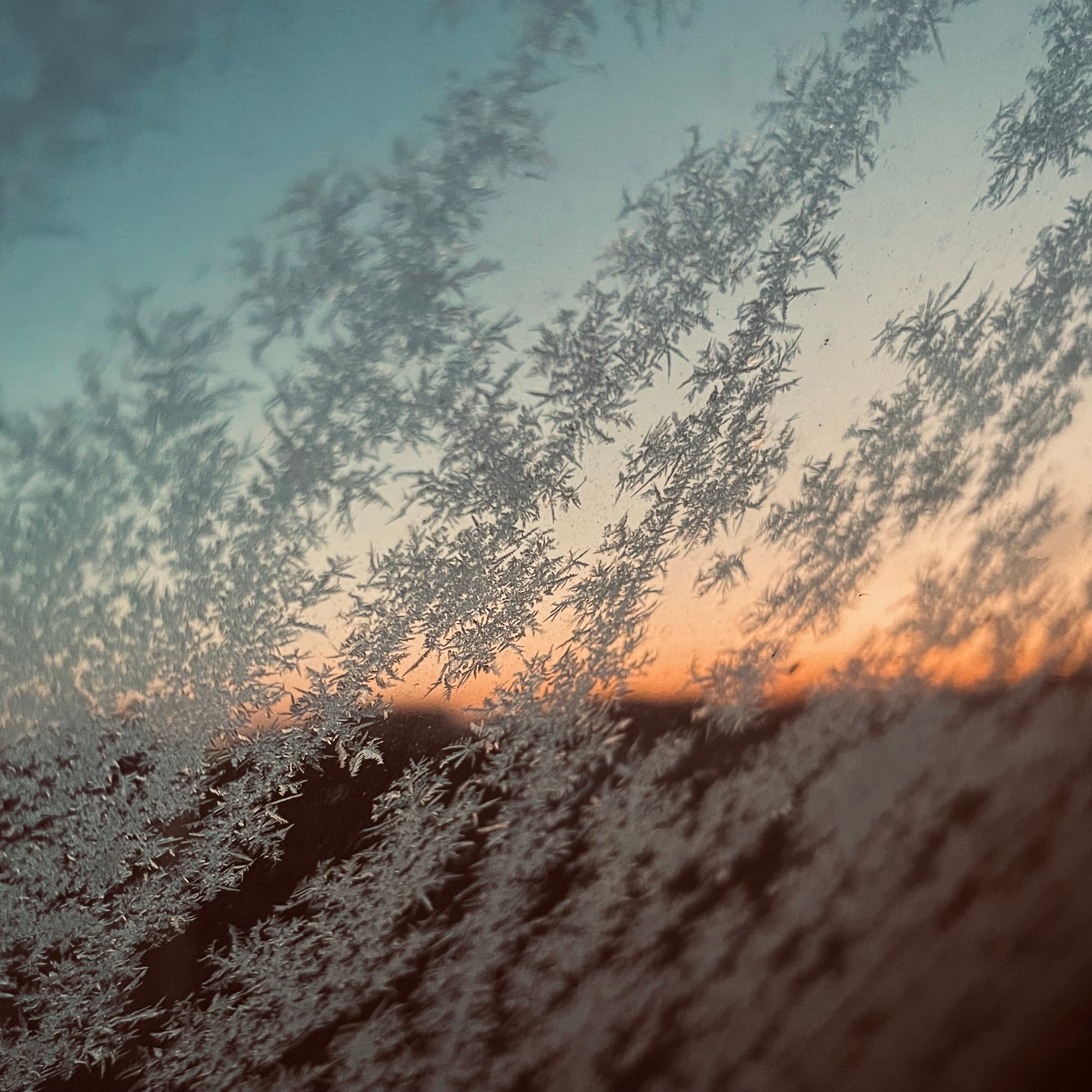 Am Fenster haben sich Eiskristalle, Eisblumen gebildet. Im Hintergrund sieht man zaghaft den Sonnenaufgang, er ist orange.