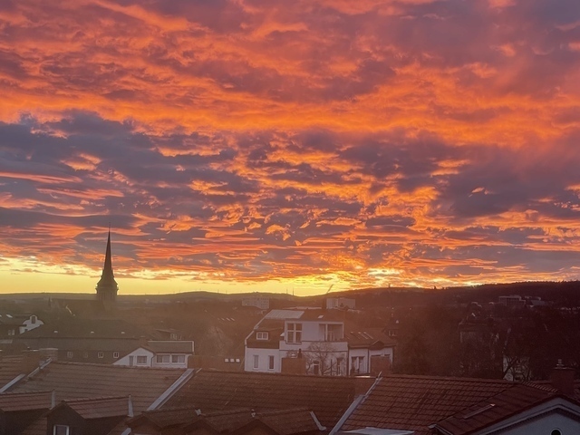 Morgenrot Schlechtwetter droht. Der rote Himmel über der Stadt, es sind Gründerzeithäuserdächer zu sehen und sehr viele Wolken. Wie Watte werden sie rot angestrahlt. Links ragt eine Kirchturmspitze in die Szenerie.