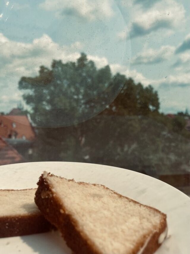 Ein Stück Zitronenkuchen liegt auf einem Teller, durch das Fenster blickt man hinaus in die Welt, Wolken und Himmel im Wechsel.