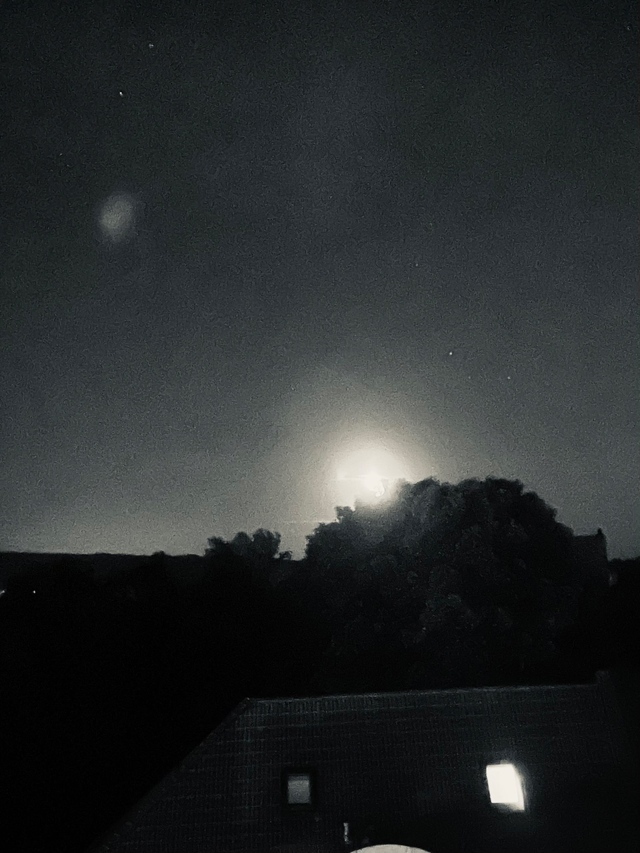 Schwarz-weiß Fotografie einer nächtlichen Szene. Über den dunklen Silhouetten von Bäumen geht ein heller Vollmond auf, dessen Licht hinter den Ästen strahlt. Am unteren Bildrand sind die Dächer eines Hauses mit einem erleuchteten Fenster sichtbar. Der Nachthimmel ist dunkel, mit wenigen kleinen Sternen und einem leichten Lichtreflex.