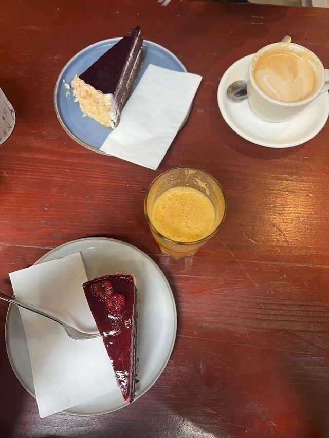 Ein Holztisch mit Tellern, auf diesen liegen Kuchenstücke, rot und Schoko und auch Kaffee der Crema hat.
