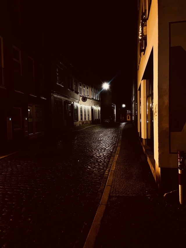Eine sperlich beleuchtete Gasse in der Geschicht Stadt. Die Straße besteht aus Kopfsteinpflaster, links und rechts befinden sich Gehwege. Es leuchten Lichter.