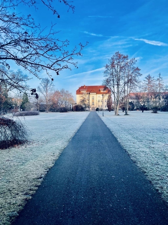 Ein Weg teilt eine Rasenfläche, links und rechts. Am Ende steht ein großes Haus mit rotem Dach. Der Himmel ist blau. Der Rasen ist vereist.